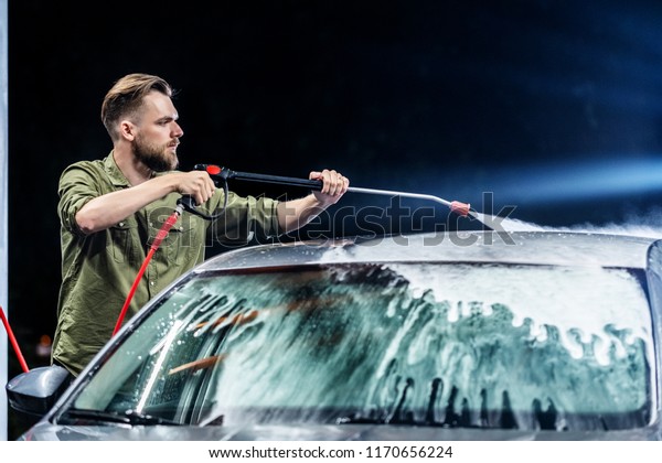 A man with a beard washes a gray car with a\
high-pressure apparatus at night in a car wash. Expensive\
advertising photography