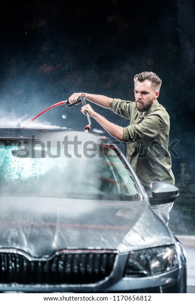 A man with a beard washes a gray car with a
high-pressure apparatus at night in a car wash. Expensive
advertising photography