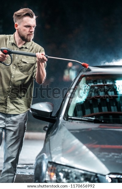 A man with a beard\
washes a gray car with a high - pressure washer in a car wash at\
night with studio light