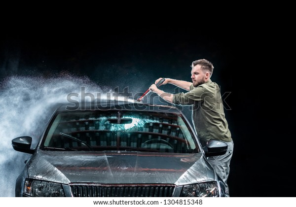 A\
man with a beard washes a gray car with a high-pressure washer at\
night in a car wash. Expensive advertising\
photography