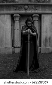 Man with a beard wearing a black hooded cape. He is a knight holding a large sword on the ground. Grave crypt in the background. Black and white.