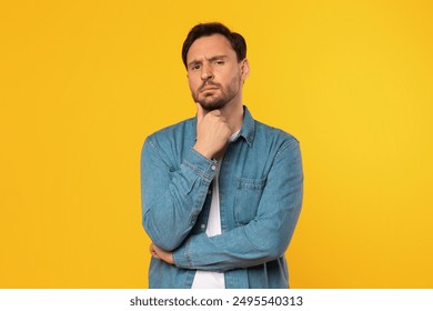 Man with a beard, wearing a blue denim shirt, stands in front of a yellow background, looking directly at the camera with a thoughtful expression. He has his hand on his chin and his arms are crossed - Powered by Shutterstock