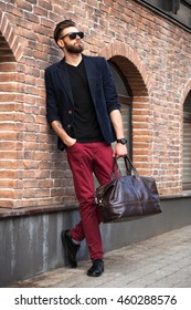 A Man With A Beard , Wearing Glasses, Wearing A Jacket In His Hand Bag For Travel On The Background Of A Brick Wall In The City.