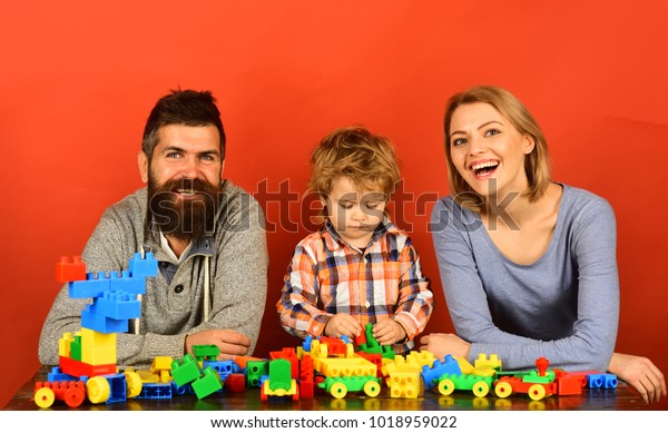 Man with beard, woman and boy play on red\
background. Family with happy and busy faces build toy cars out of\
colored construction blocks. Parents and kid in playroom. Childhood\
and playing concept.