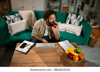 Man with a beard working remotely on a laptop, sitting on a sofa and enjoying a healthy apple snack at home