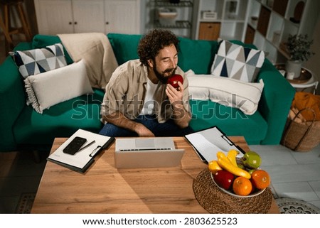 Man with a beard working remotely on a laptop, sitting on a sofa and enjoying a healthy apple snack at home