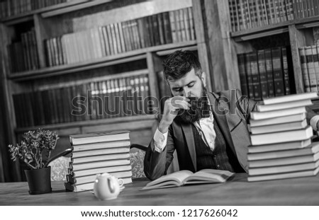 Man or bearded teacher reads book. Research and studying concept. Man in classic suit, professor with concentrated busy face sits in library near piles of books, bookshelves on background, defocused.