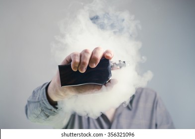A man behind a cloud of vapor holds up an e-cigarette mod. 