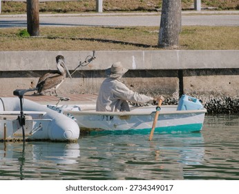 Man in Beige Hat Rows Small Boat as Pelican Perches Near Dockside Waterfront.  Calm water while a pelican rests on the dock, creating a tranquil harbor scene with nautical charm and nature meeting craft.
