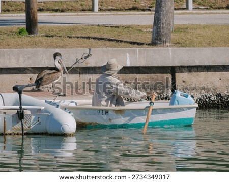 Man in Beige Hat Rows Small Boat as Pelican Perches Near Dockside Waterfront.  Calm water while a pelican rests on the dock, creating a tranquil harbor scene with nautical charm and nature meeting craft.
