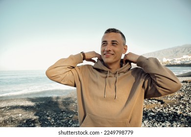 A man in a beige hoodie against the background of the sea. A guy is walking on the beach and enjoying the views, happiness from traveling in the summer, freedom. - Powered by Shutterstock