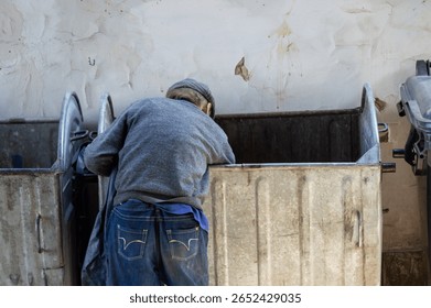 A man bends over two trash bins in an alley, looking for items of value under the warm afternoon sunlight