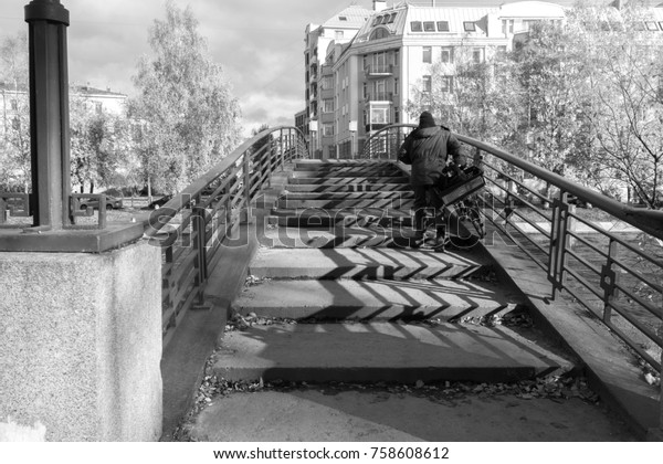 Man with bicycle climbs up the steps on\
the bridge over the river. View of the city, street with parked\
cars and historic buildings. Black and white\
color.