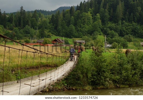 Man with\
bicycle crossing old suspension bridge over the mountain river.\
Beautiful rustic landscape view on Carpathian hills. Ukrainian\
village. Green tourism\
concept.