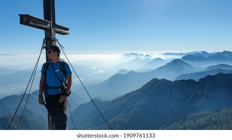 A man with big backpack standing under a metal cross with a panoramic view on the Alps from the top of Mittagskogel in Austrian Alps. Clear and sunny day. A bit of haze in the valley. Outdoor activity