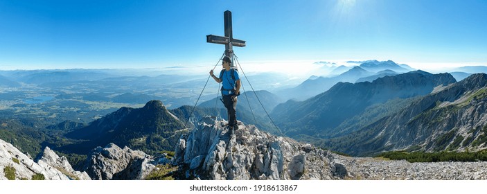 A man with big backpack standing under a metal cross with a panoramic view on the Alps from the top of Mittagskogel in Austrian Alps. Clear and sunny day. A bit of haze in the valley. Outdoor activity