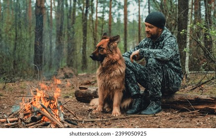 Man with big dog sits at the fire in the forest. Human looks at the fire and touching his dog in nature in autumn. Alone traveler is resting with his German shepherd by the forest fire. - Powered by Shutterstock