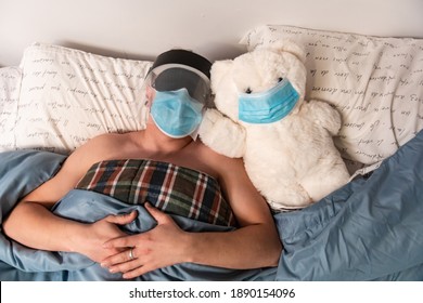 A Man And A Big White Teddy Bear In Bed, Both Wearing Coronavirus Protective Mask. Mas Also Wars Sleeping Mask And Plastic Visor. Ironic.