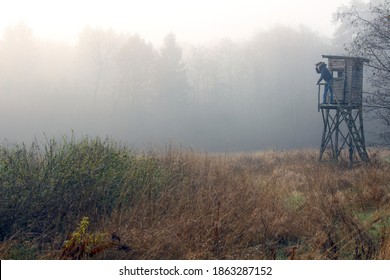 Man with binoculars on raised hide in foggy fields looking out for animals. Ranger and biologist in wilderness. Germany during winter winter morning.