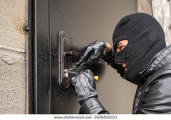 A man in a black balaclava mask opens a\
locked door with a lock pick. The robber breaks into the house.\
Robbery of a private house. Criminal\
concept