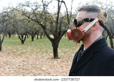 A man in a black coat, business suit, gas mask and steampunk goggles poses in an autumn park. Horizontal photo
