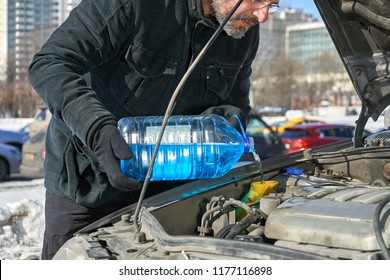 Man In Black Coat Filling A Windshield Washer Tank Of A Car With A Wiper Fluid Standing On The Roadside Of Busy Moscow Street In Winter Day                              