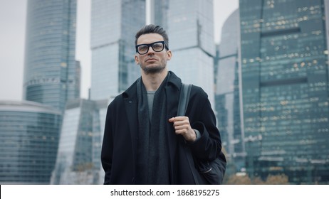 Man In Black Coat Slow Motion Walking On Background Of Business City Skyscrapers In The Evening, Lights In The Windows. Gimbal Shot Of Young Businessman In Eyeglasses