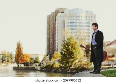 A Man In A Black Coat Walks With A Guitar Case In The City Along The River.