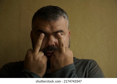 Man With Black Eyes Wearing Shirt Standing Over Wallpaper Background. Showing Two 2 Middle Fingers, Impolite And Rude. Expression Sign, Symbol. Fingers In Front Of The Pupils Of The Eyes