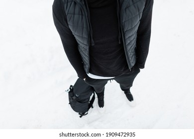 Man In Black Fashionable Clothes With A Hoodie, Vest, Pants, Sneakers And A Backpack Stands In The Snow. Mens Black Winter Style 