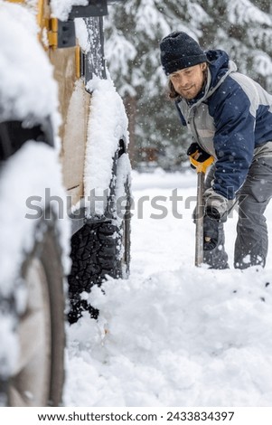 A man in a black hat and blue jacket is shoveling snow off the back of a yellow jeep