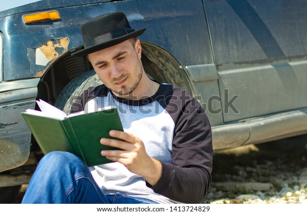 A man in a black hat read a book near the car\
in a mountains
