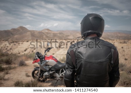 A man in black helmet and black leather jacket standing near adventure motorcycle in desert, adventure overland travel, back view