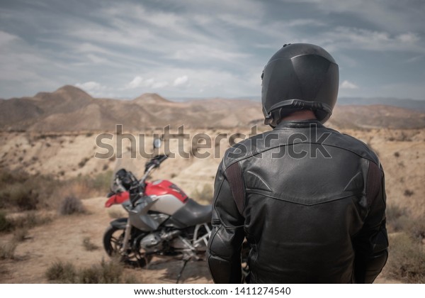 黒いヘルメットをかぶった男性と黒い革のジャケットが砂漠の冒険バイクの近くに立ち アドベンチャーオーバーランド旅行 バックビュー の写真素材 今すぐ編集