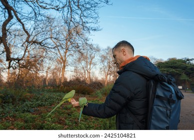 A man in a black jacket examines a large leaf during a late afternoon walk in a natural setting, surrounded by trees and fallen leaves, evoking a serene, seasonal mood in London's park. - Powered by Shutterstock