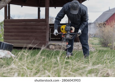 A man in a black jacket and hat uses a gas-powered grass trimmer to cut grass in a rural area. A wooden gazebo and a red barn are in the background.