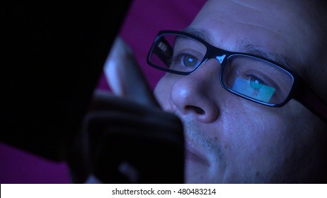 Man In Black Rim Glasses Using His Tablet Computer In Dark Room. Screen Reflection In The Lens