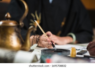 A man in a black shirt dips a brush for traditional Chinese calligraphy in ink. In the foreground is a copper kettle in defocus. on the table is paper with hieroglyphs. close-up.