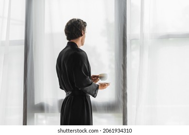 Man In Black Silk Robe Holding Cup Of Coffee Near Curtains At Home