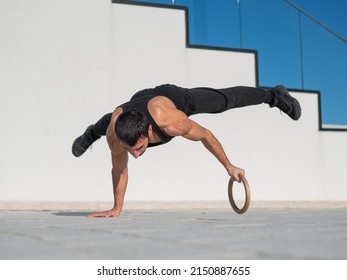 A man in black sportswear does an acrobatic handstand on a ring outdoors.