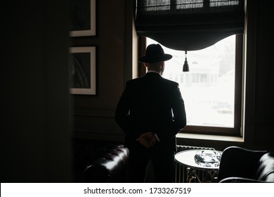 Man In A Black Suit And Hat Stands With His Back To Camera. Look Out The Window. Silhouette Photography, Film Frame. Dickens Style Man Looking Out Window, Arms Crossed Behind