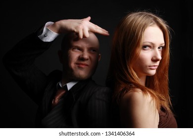 Man In Black Suit Make Finger Pistol And Shot His Woman In Dress Stand Back To Back Playing Secret Service Agent And His Lover. Studio Portrait Against Black Background