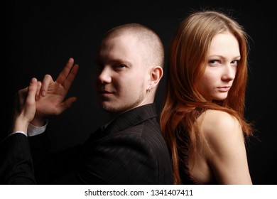 Man In Black Suit Make Finger Pistol And Woman In Dress Stand Back To Back Playing Secret Service Agent And His Lover. Studio Portrait Against Black Background