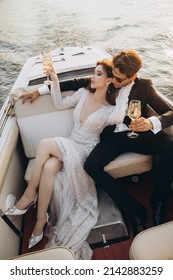 Man In A Black Suit And Sunglasses Hugs A Woman On A Yacht On A Sunny Day. Couple Drinking Champagne And Having Fun