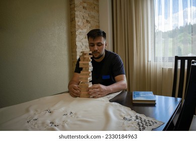 A man in a black T-shirt pulls a block from a wooden structure at a table in order to place it on top of the structure