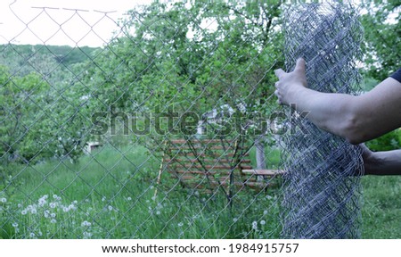 a man in a black T-shirt unfolds a galvanized netting against the background of a green garden or farm, installing a metal fence in a natural area, fencing a part of the village land with a metal net