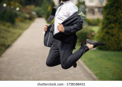 A Man In A Black Tuxedo Suit, White Shirt And Leather Shoes With A Long Tie Is Jumping In Nature On A Green Background. A Stylish Stylish Groom In A Jump Hold On To His Jacket With His Hands. 