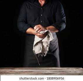 Man in black uniform holds gray linen rag and wipes his hands, chef stands on black background, close up