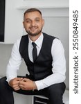 A man in a black vest and white shirt is smiling. He is wearing a black tie and a silver bracelet