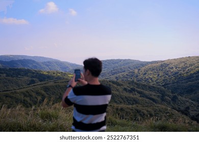 Man in black and white stripes, back facing with phone, admiring mountain view with green hills in sunlight under blue cloudy sky. Nature, landscape, outdoor adventure, peaceful, serene, mountains. - Powered by Shutterstock